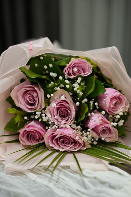 A bouquet of fresh, pastel pink roses with tightly rolled petals and vibrant green leaves, accented with delicate white baby's breath flowers. The flowers are arranged in a circular, compact bouquet a