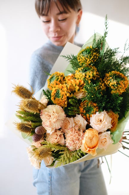 A young woman holding a large, vibrant bouquet of fresh flowers in a variety of colors and textures. The arrangement includes peach and cream roses, bright yellow and orange marigolds, and green folia
