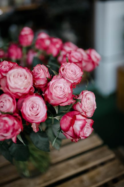 A vibrant bouquet of pink roses with layered, ruffled petals and green stems, arranged in a clear glass vase. The roses are fresh, showing a glossy surface and firm petals, with some buds still partia