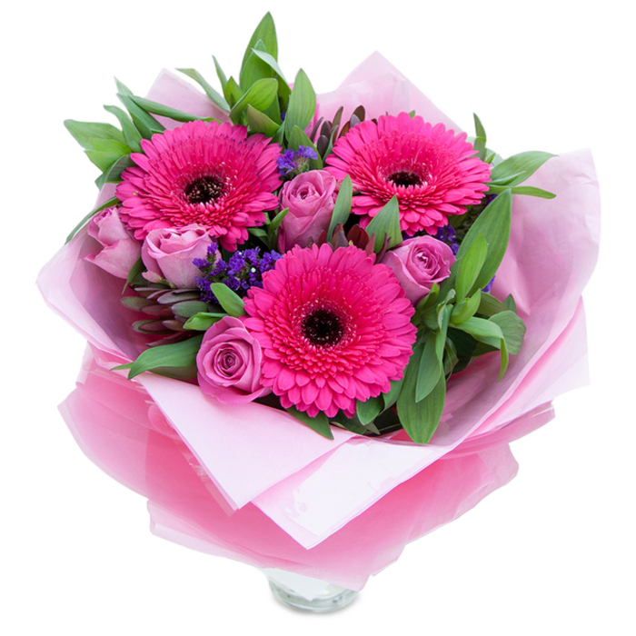 I see a bouquet that balances bold cheer with gentle grace: three vibrant fuchsia Gerbera daisies fan out with confident petals and dark, almost inky centres, anchoring the arrangement like joyful faces. Around them, roses in a soft lavender-pink or dusty rose are carefully placed, their tightly rolled petals suggesting freshness and a slow, unfolding sweetness. Sprigs of tiny purple statice float between the larger flowers, adding tiny islands of texture and a cooler hue that makes the pinks sing. A generous background of green foliage-broad salal or eucalyptus-frames the blooms with crisp, slightly glossy leaves, contributing a faintly herbal aroma that complements the roses' soft scent. For presentation, the bouquet is wrapped in an inner blush-pink matte tissue that cushions the stems, then enveloped in a sheer fuchsia-pink cellophane with a gentle crinkle that catches the light and deepens the pink theme. The lower part reveals the top rim of a clear glass vase, a quiet promise that the arrangement is ready to stand in water and brighten a room. The lighting is soft and even, accentuating petal textures and the velvet-like swirl of the rosebuds. This composition feels made for small celebrations-birthdays on the village green, a Mother's Day surprise after a walk along the Grand Union Canal towpath, or an anniversary table centerpiece-and carries the kind of careful hand that local customers in and around Kings Langley have come to expect from their florist.